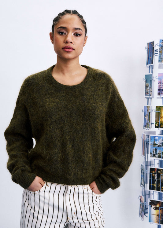 A woman wearing a fuzzy dark olive green crewneck sweater and white pants with thin black vertical stripes, standing against a white wall with a poster rack on the right.